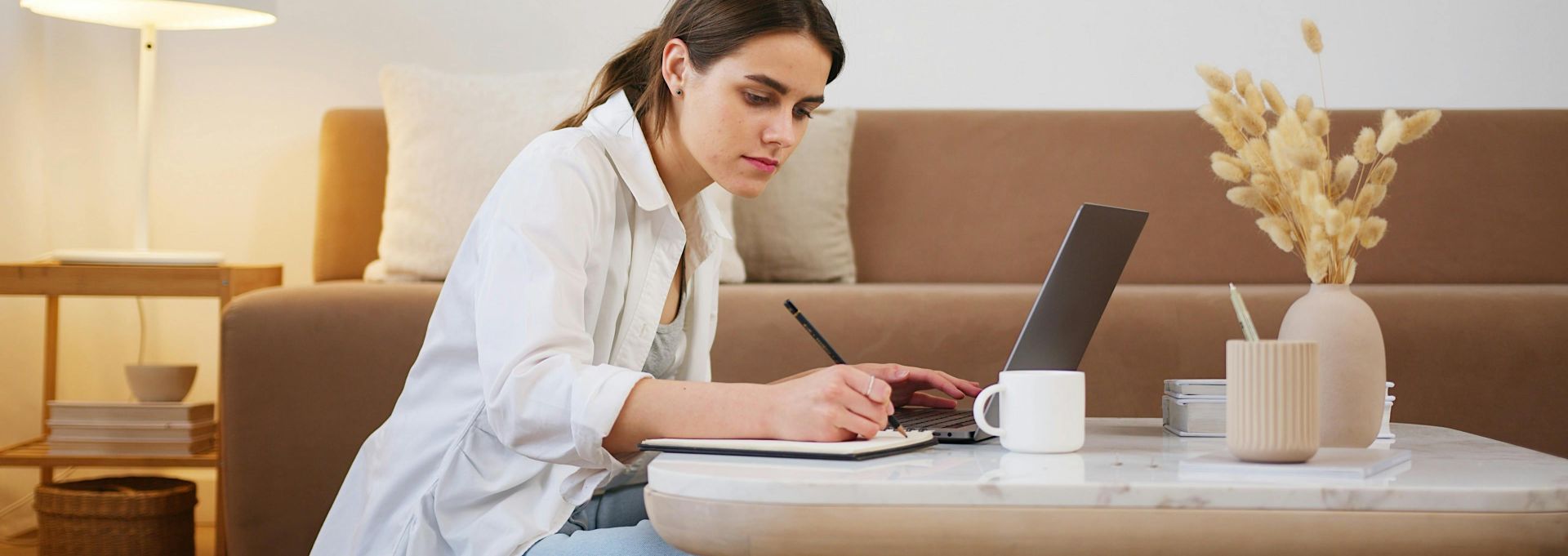 Side view full length young focused female in casual clothes browsing modern laptop and writing down notes while sitting on floor in cozy modern apartment